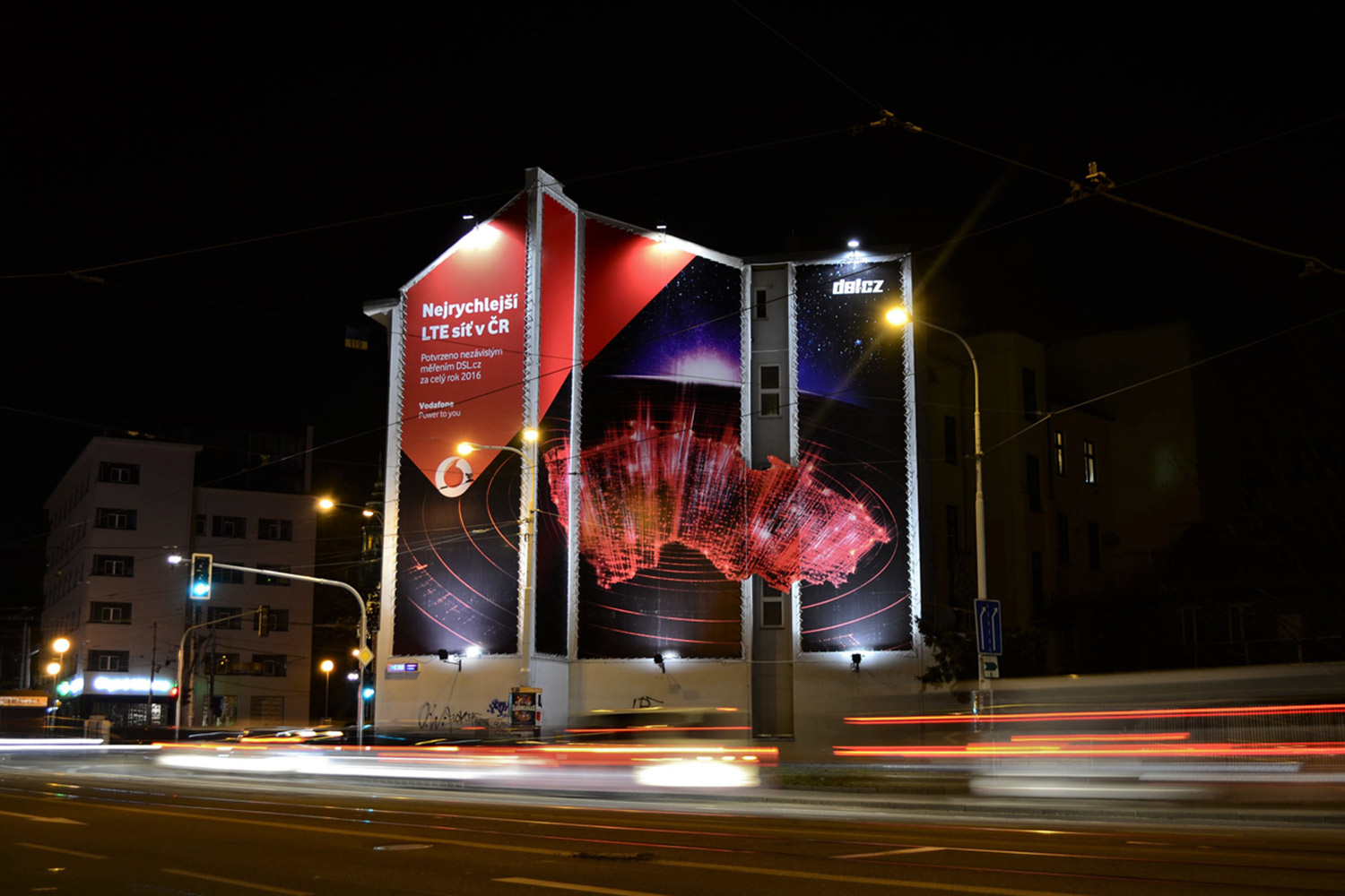 Reklamní plocha na štítu domu na Mendlově náměstí v Brně. Reklamní kampaň společnosti Vodafone. Noční snímek s dlouhým časem. Rozmazaná světla.