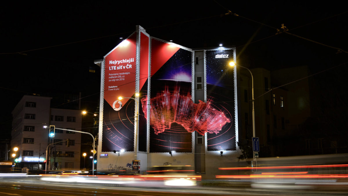 Reklamní plocha na štítu domu na Mendlově náměstí v Brně. Reklamní kampaň společnosti Vodafone. Noční snímek s dlouhým časem. Rozmazaná světla.