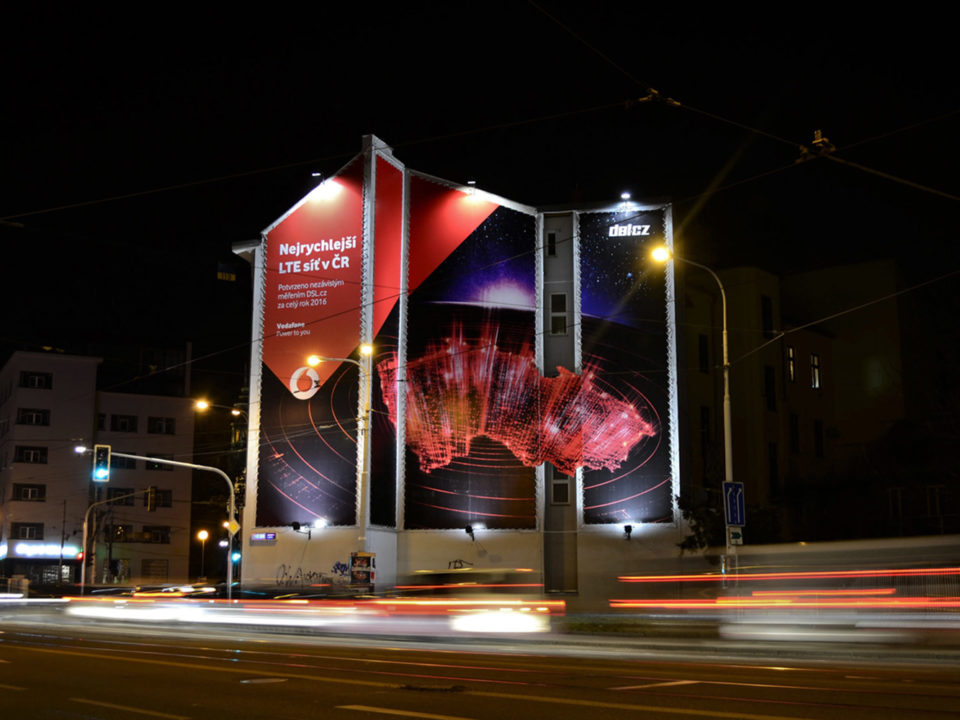 Reklamní plocha na štítu domu na Mendlově náměstí v Brně. Reklamní kampaň společnosti Vodafone. Noční snímek s dlouhým časem. Rozmazaná světla.